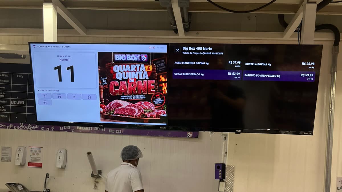 Balcão do açougue do Big Box 408 Norte com o SuaVez: à esquerda a TV 1 com a senha atual 11 e uma campanha da loja, à direita a TV 2 com a tabela de preços, e na parede o dispositivo de controle roxo do açougueiro